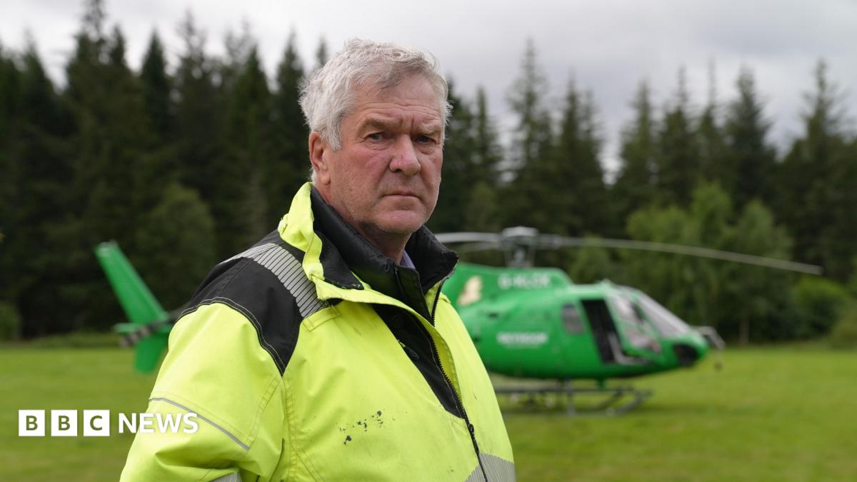 Nethy Bridge community council leader Sandy McCook wearing a high-visibility yellow jacket stands in a grassy clearing with a green helicopter parked behind them. Dense conifer forest forms the background under an overcast sky.