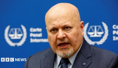 International Criminal Court Prosecutor Karim Khan speaks during an interview while sitting in front of a blue sign with blurry white writing and the symbol of the ICC - the scales of justice surrounded by a laurel wreath.