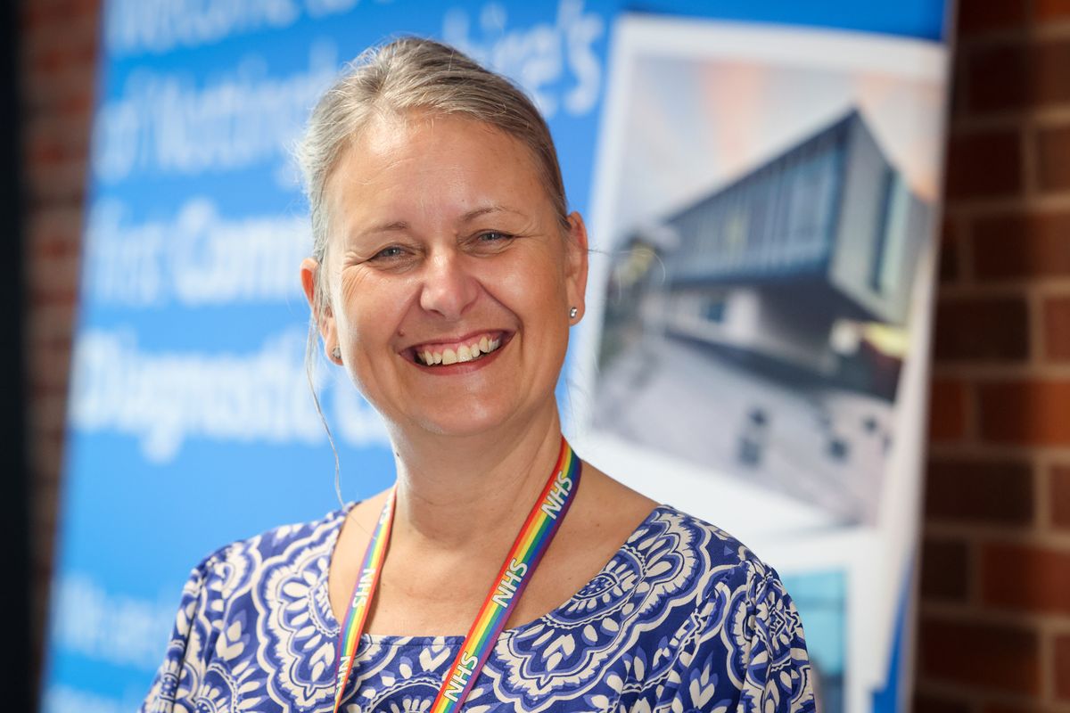 Middle-aged woman indoors with blue and white patterned blouse and rainbow NHS lanyard on, smiling at camera in front of blurred blue/white NHS banner poster in background and brick wall