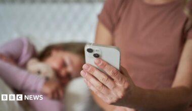 A mum, whose face cannot be seen, holds a smartphone out in front of her while her young daughter lies in bed beside her. The child's face is blurred.