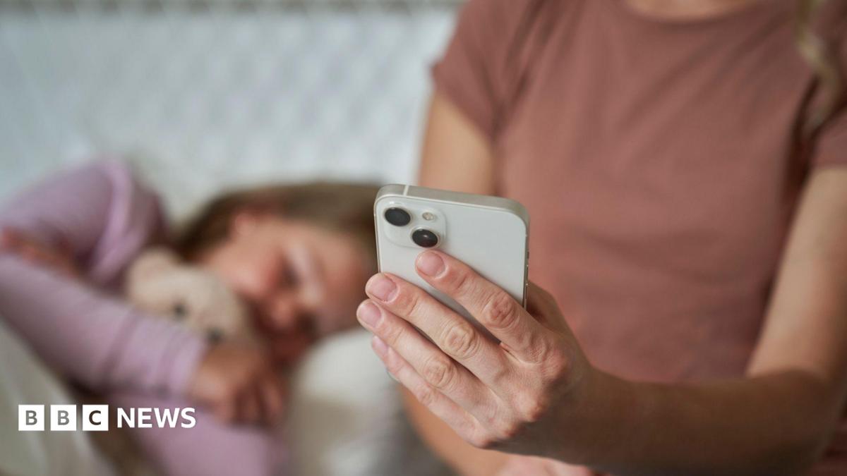 A mum, whose face cannot be seen, holds a smartphone out in front of her while her young daughter lies in bed beside her. The child's face is blurred.