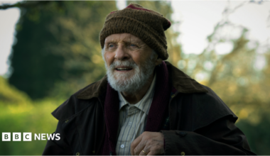 Daniel wears a blue cap and blue jacket with blue, red and green stripes on his arms. He rests his head on Jack's shoulder. Jack rests his hand on Daniel's head and his face in his head. Jack wears a blue wool hat and blue waterproof coat.