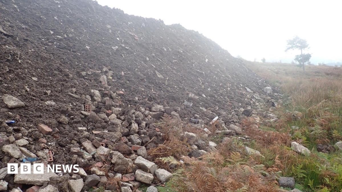 A large pile of rubble and dirt has been dumped in the middle of a grassy field. A tree can be seen in the distance.