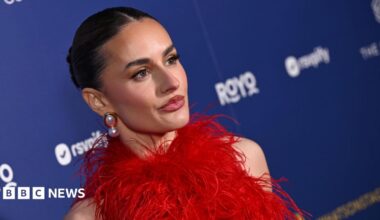 A zoomed in, slanted angle of Amber Davies, in a red feathered dress and wearing big pearl earrings. She stands in front of a branded blue wall with sponsor logos. Her dark hair is pulled back into a slick bun.