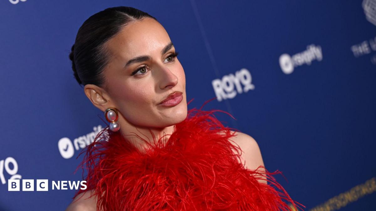 A zoomed in, slanted angle of Amber Davies, in a red feathered dress and wearing big pearl earrings. She stands in front of a branded blue wall with sponsor logos. Her dark hair is pulled back into a slick bun.