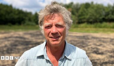 Guy Singh-Watson is wearing a blue short-sleeved shirt as he stands in the bright sunshine in a dry reservoir on a farm at Riverford Organic Farmers in Devon.