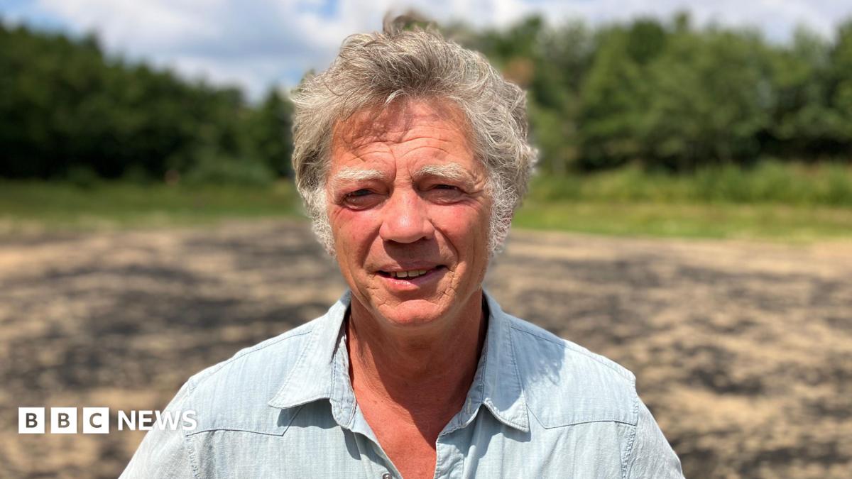 Guy Singh-Watson is wearing a blue short-sleeved shirt as he stands in the bright sunshine in a dry reservoir on a farm at Riverford Organic Farmers in Devon.
