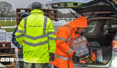 A man in a yellow hi-vis jacket with the word South East Water on the back. Another man, in an orange hi-vis jacket, is carrying a slab of water out of a car.