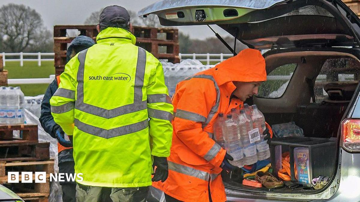 A man in a yellow hi-vis jacket with the word South East Water on the back. Another man, in an orange hi-vis jacket, is carrying a slab of water out of a car.