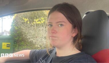 Beatrix sat in the driver's seat of her car, looking at the passenger who is taking a photo of her with a slightly grumpy expression on her face. She has shoulder-length black hair and is wearing a black T-shirt with a purple pattern on the front.