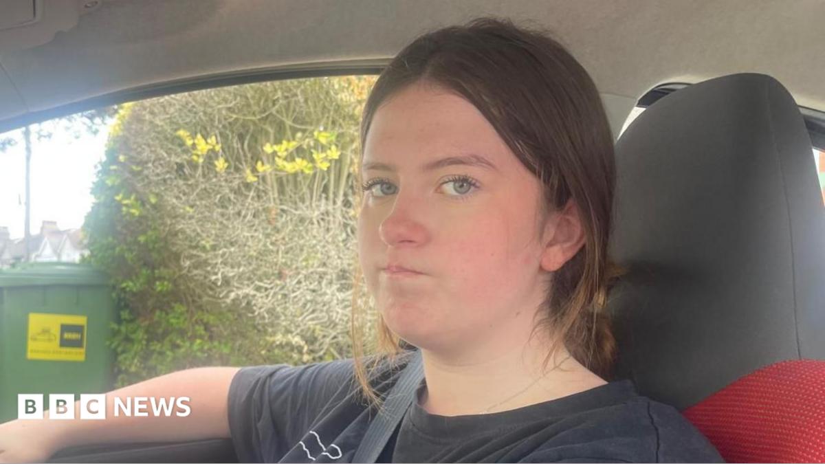 Beatrix sat in the driver's seat of her car, looking at the passenger who is taking a photo of her with a slightly grumpy expression on her face. She has shoulder-length black hair and is wearing a black T-shirt with a purple pattern on the front.