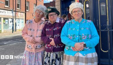 Three women are standing together on a high street wearing colourful knitted cardigans with their hands folded on top of their stomach. They are all wearing glasses and have grey wigs on their heads.