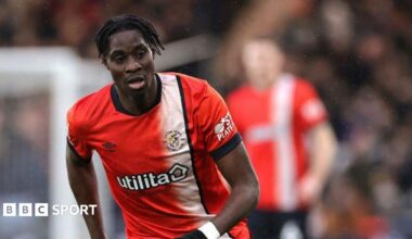 Elijah Adebayo in match action for Luton wearing an orange playing shirt with a white vertical stripe from the left shoulder down the front of the shirt