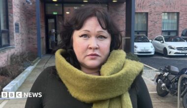 Sara Morrison with short dark hair looks at the camera. She is wearing a pale green scarf and is standing outside in front of a building and a number of parked cars.