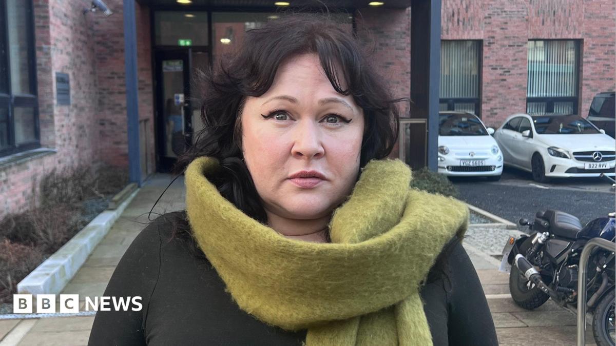 Sara Morrison with short dark hair looks at the camera. She is wearing a pale green scarf and is standing outside in front of a building and a number of parked cars.