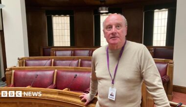 Rob Roche, is standing in the main chamber at Luton Borough Council. He is wearing a beige jumper and is stood in front of a number of red seats.
