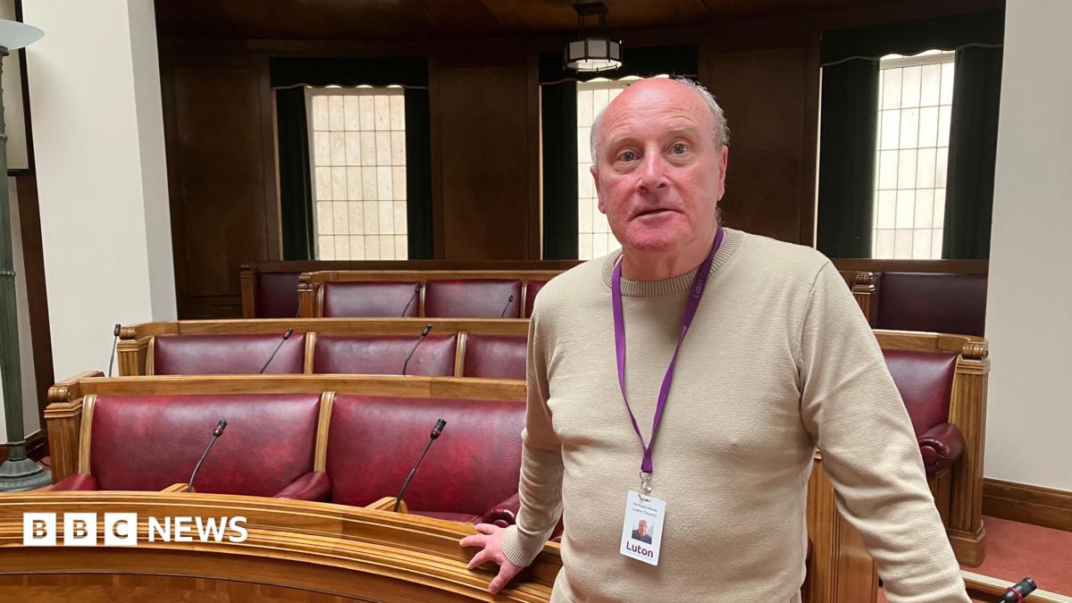 Rob Roche, is standing in the main chamber at Luton Borough Council. He is wearing a beige jumper and is stood in front of a number of red seats.