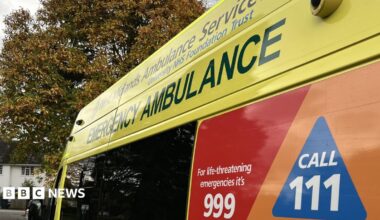 A close-up shot of the side of an ambulance. At the top it says West Midlands Ambulance Service University NHS Foundation Trust. Below that in larger lettering it says emergency ambulance, and at the bottom in white lettering against a red background it says for life-threatening emergencies it's 999