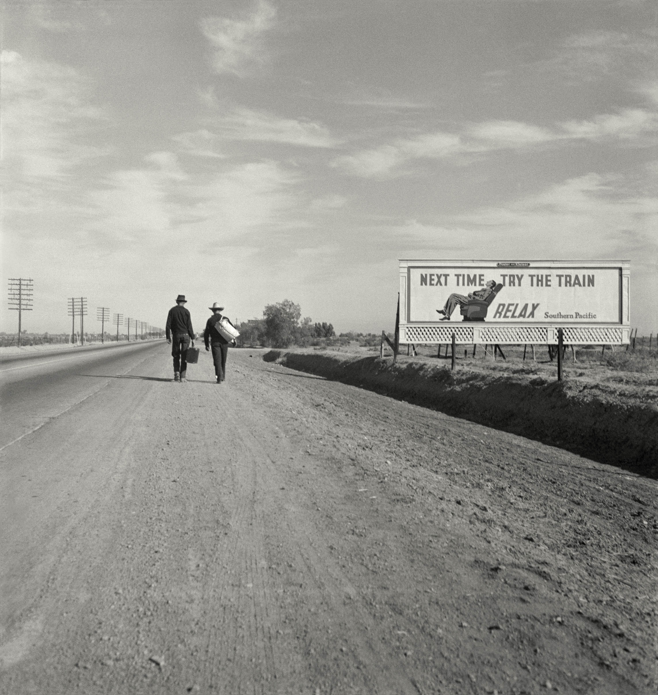 Two men carrying luggage walk away from the camera along a flat California highway, passing a billboard advertising Southern Pacific railroad with the words "Next time try the train — relax."