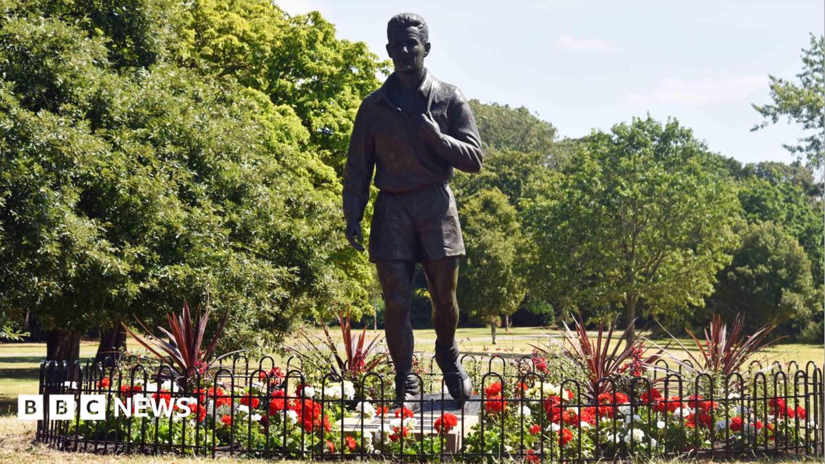 A life-size, grey sculpture of Brian Clough. He appears to be walking with his football boots slung over his shoulder by their laces. The figure has short hair and is wearing shorts and a long sleeve top. The statue is surrounded by red and white flowers and there are trees in the background.