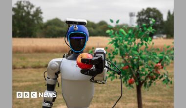 A humanoid robot holds a red apple in the air it has just picked from a tree.