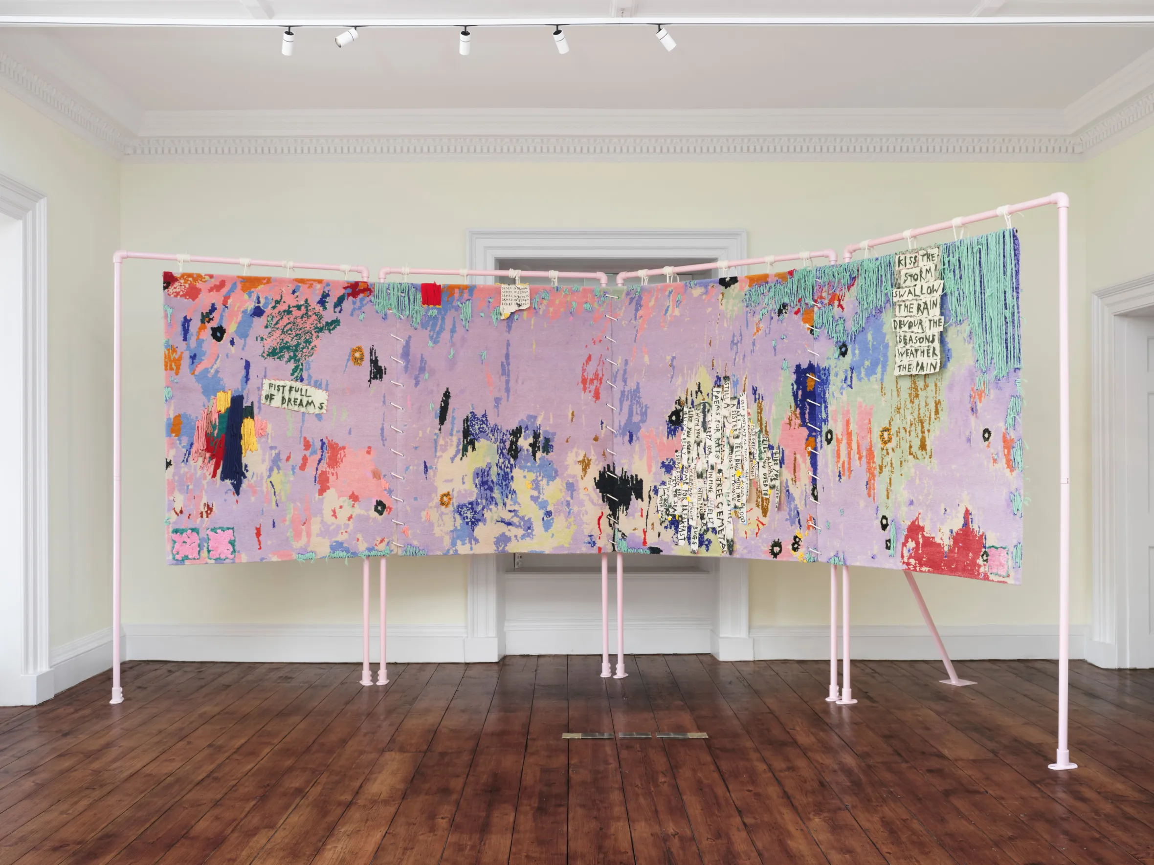 Installation view of Daisy Parris's "Fist Full of Dreams," a vibrant textile piece with abstract patterns, text, and fringe, displayed on a pink frame.