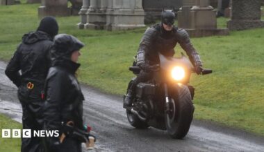 Filming on a scene in The Batman - a man dressed in a Batman costume is riding a motorbike in a graveyard, watched by crew members