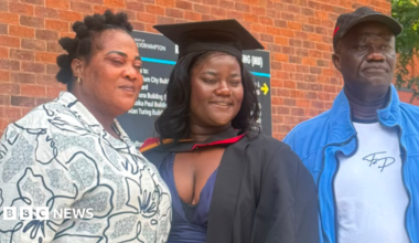 A woman in the middle is wearing a black graduation gown. Another woman on the left is wearing a dark and white top, while a man on the right is in a dark baseball cap and blue and white clothing. A building featuring a sign is behind them.
