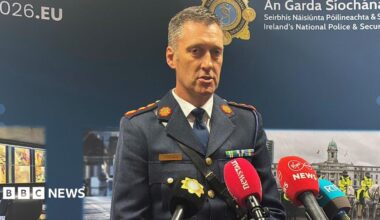 Garda officer wearing a blue uniform. He has short dark and grey hair and has mics in front of him. An Garda Siochana is on the background behind with police pictures.