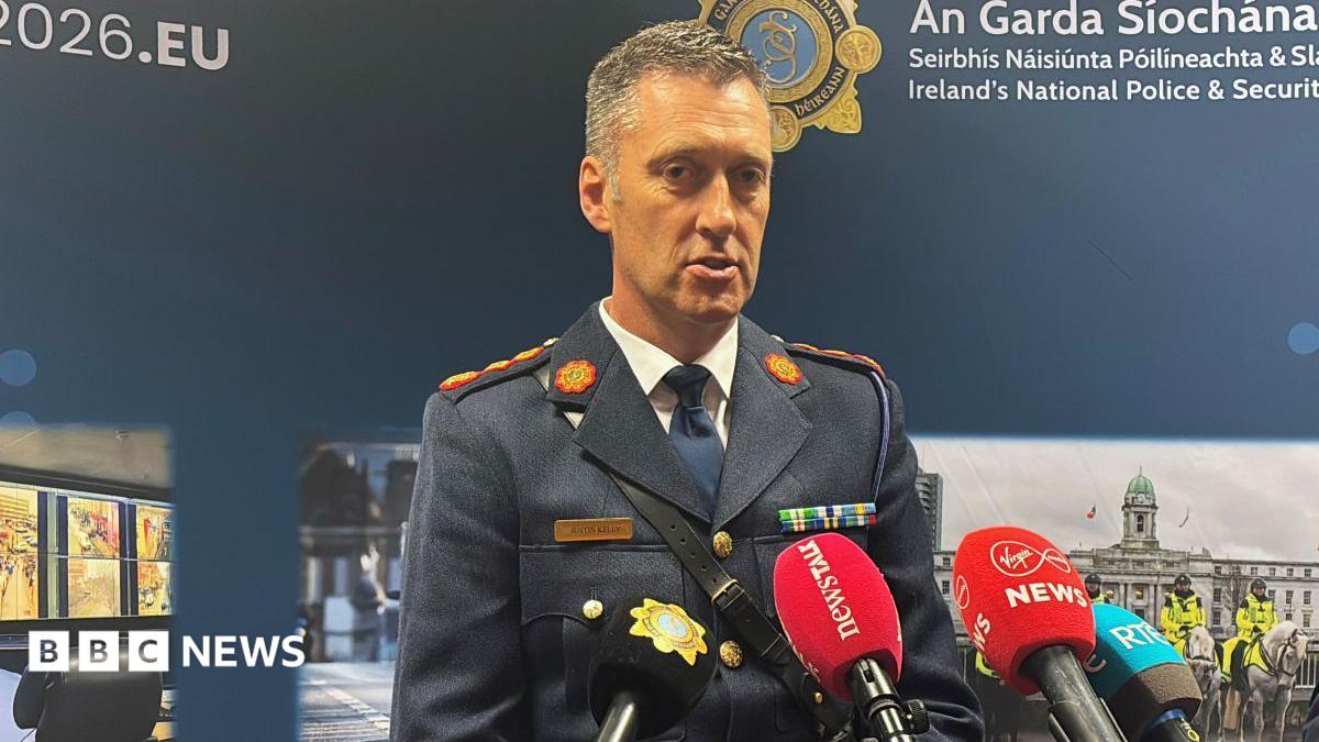 Garda officer wearing a blue uniform. He has short dark and grey hair and has mics in front of him. An Garda Siochana is on the background behind with police pictures.