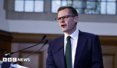 Reform UK's Robert Jenrick mid-speech. He is wearing glasses, has short, dark hair and is wearing a collared shirt and suit with a green tie.