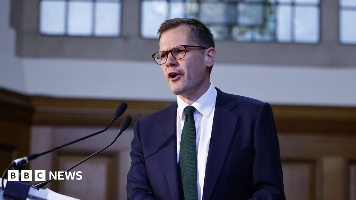 Reform UK's Robert Jenrick mid-speech. He is wearing glasses, has short, dark hair and is wearing a collared shirt and suit with a green tie.
