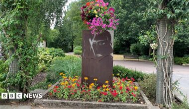 The new artwork, called "XhibitA" in Queen's park in Swindon. The artwork sits in a square flower bed with yellow and red flowers at is base. Facing the camera is a recreation of XTC's 1979 "Drums and Wires".