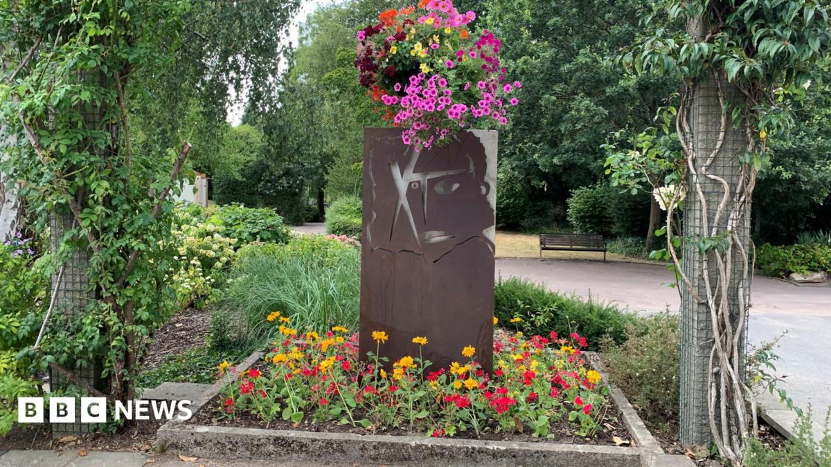 The new artwork, called "XhibitA" in Queen's park in Swindon. The artwork sits in a square flower bed with yellow and red flowers at is base. Facing the camera is a recreation of XTC's 1979 "Drums and Wires".