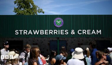A crowd of people walk past a green building. The structure has a large sign that reads "strawberries and cream" and has the Wimbledon tennis logo.