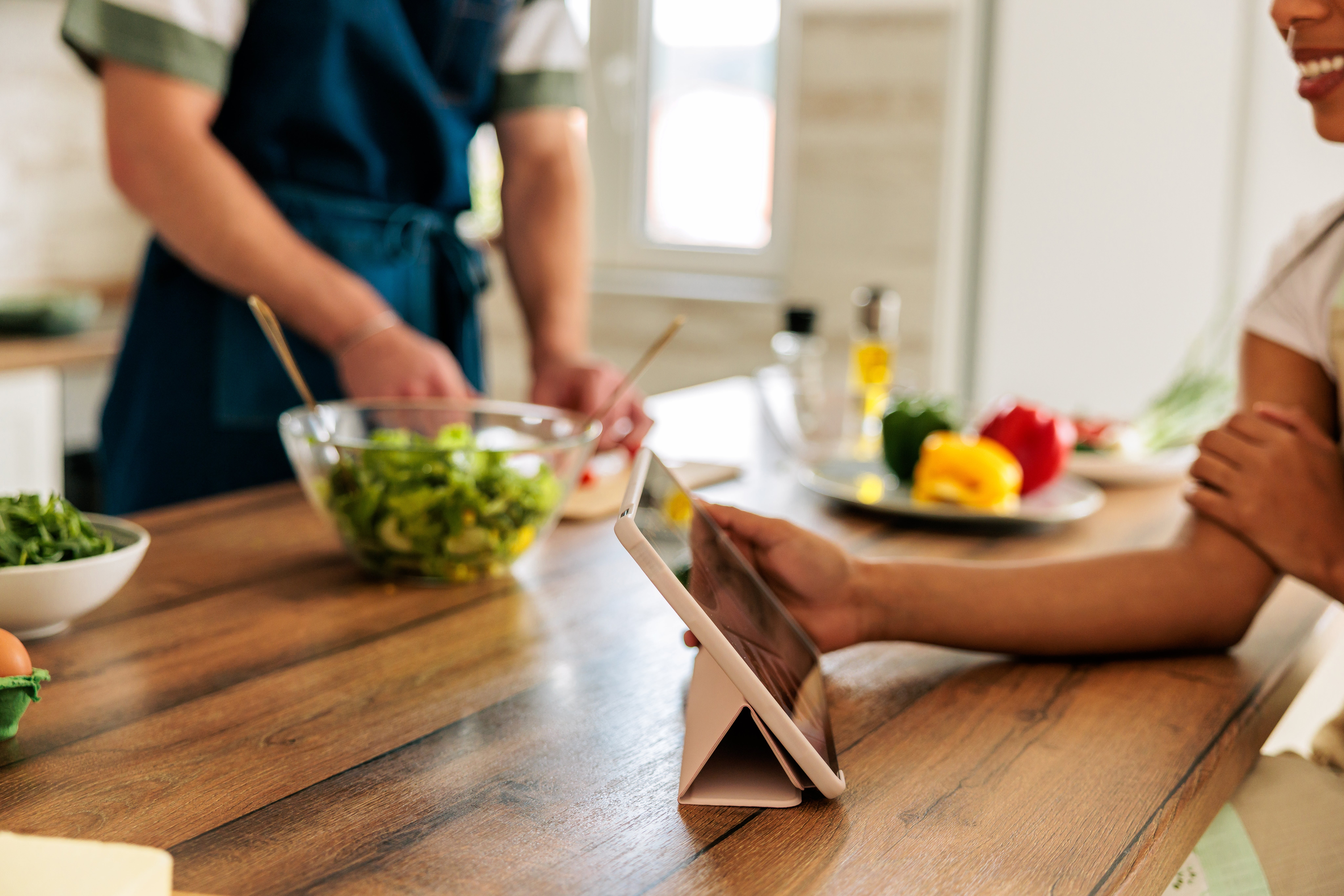 Person preparing salad in a modern kitchen, another person viewing a tablet, suggesting a focus on cooking or meal planning