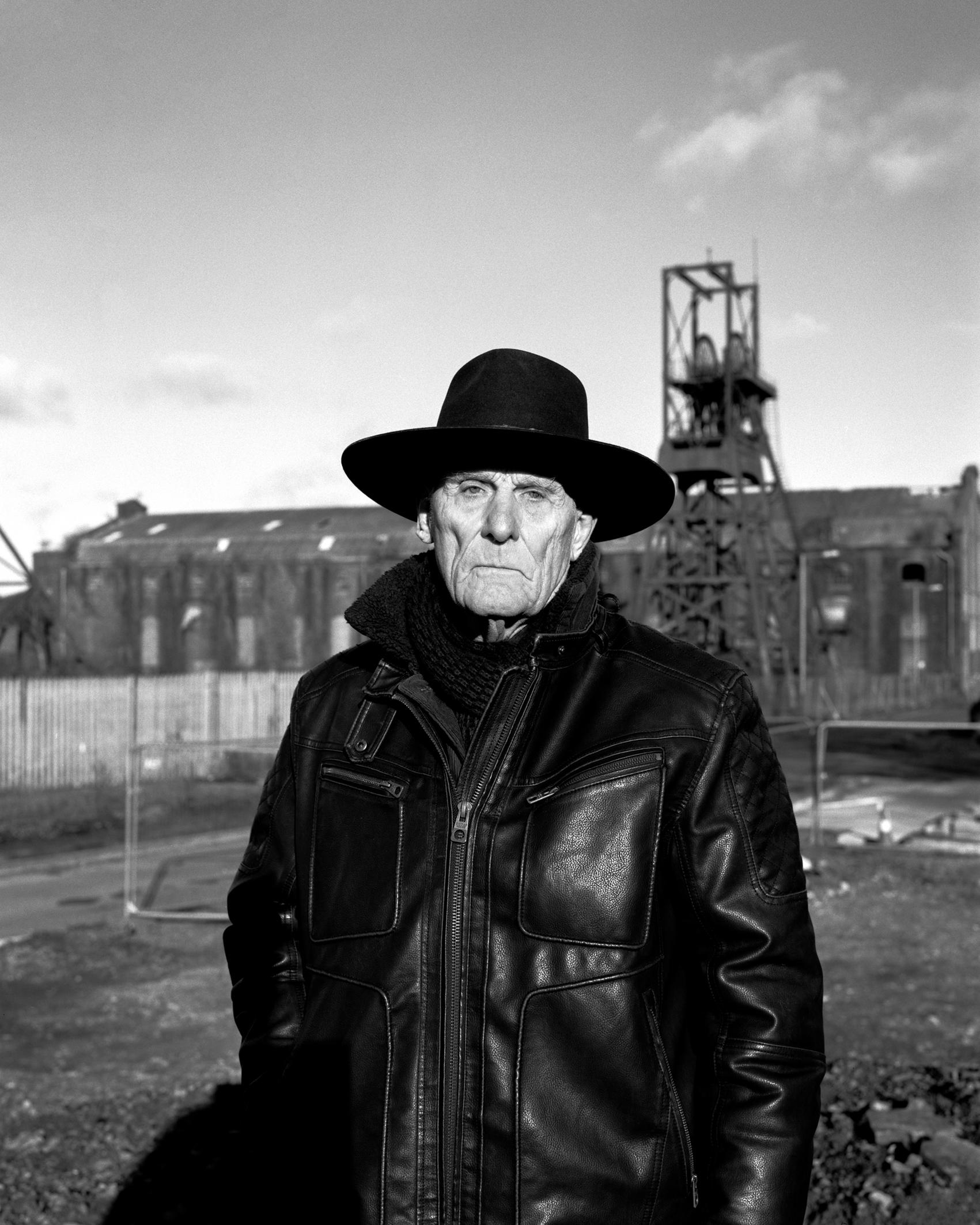 Black and white photo of an elderly man in a wide-brimmed hat and leather jacket standing in an industrial area with old buildings and machinery