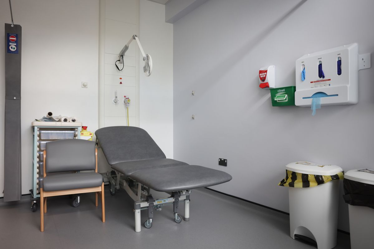 Clinical room in a hospital, with grey chair, grey reclining bed, waste bins, glove dispenser units and other medical facilities visible against white wall backdrop