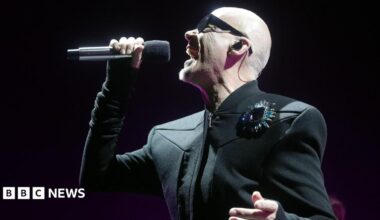 The Human League's Philip Oakey is wearing a black jacket, sunglasses and is singing into a microphone.