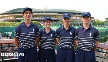 A group of four children wearing blue polo shirts with white hoops on the bottom. They have their arms behind their back and are smiling. They are stood in front of a stadium, with tennis courts also behind them.