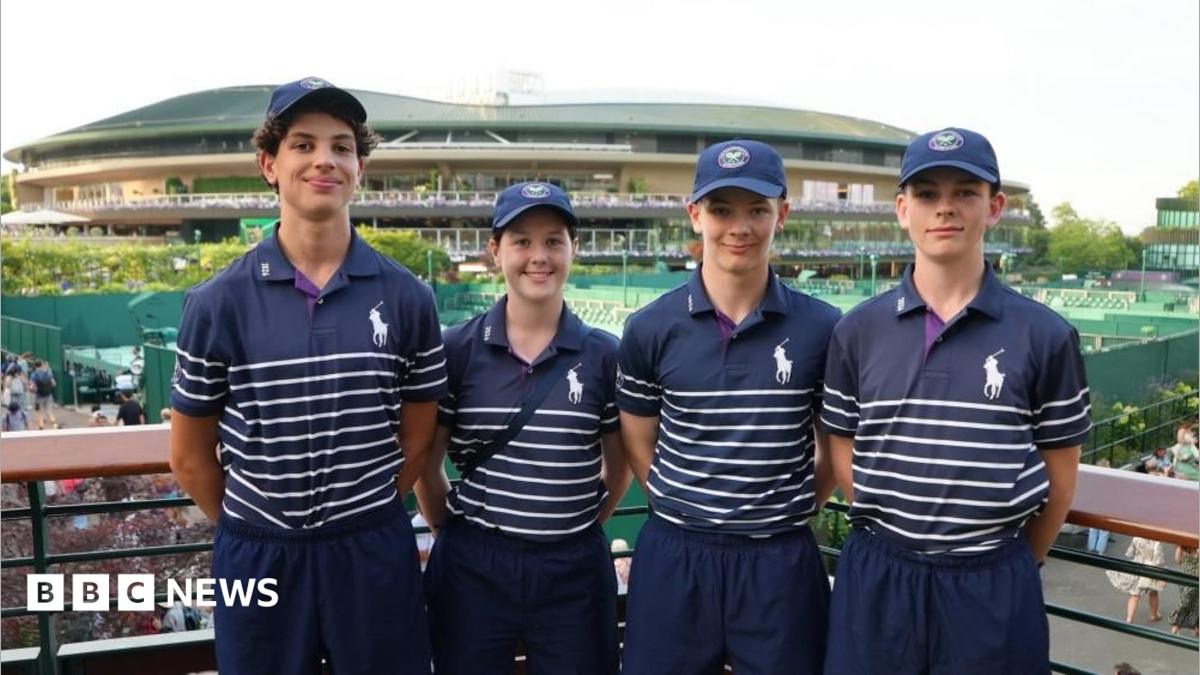 A group of four children wearing blue polo shirts with white hoops on the bottom. They have their arms behind their back and are smiling. They are stood in front of a stadium, with tennis courts also behind them.