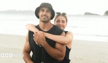 An image of a man and a woman standing on a beach. A blonde woman with sunglasses on her head has her arms around a man wearing a black vest and black bucket hat. They are both smiling at the camera.