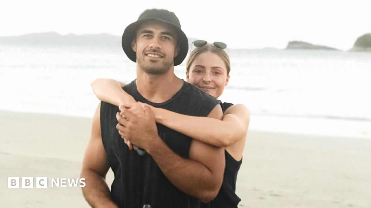 An image of a man and a woman standing on a beach. A blonde woman with sunglasses on her head has her arms around a man wearing a black vest and black bucket hat. They are both smiling at the camera.