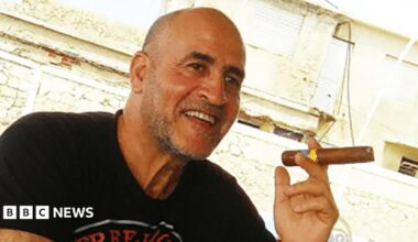 Daniel Siad, a man with a shaved head and stubble, wearing a black T-shirt, sits outside in what appears to be a sunny street, smiling with a large cigar raised in his hand.