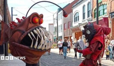 The "Humber Monster" - a person wearing a large, brown angler fish costume with bulging red eyes and sharp white teeth, confronts a person wearing a red diving suit and black diving helmet, while standing in a pedestrian high street as people watch.