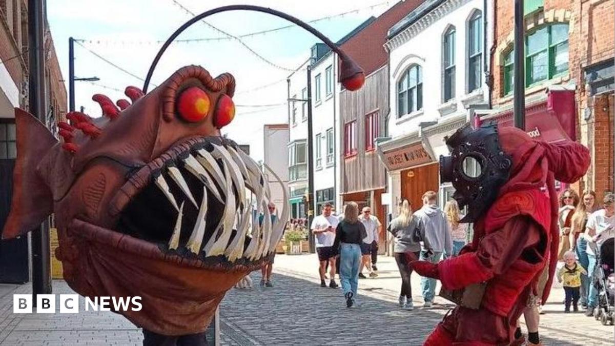The "Humber Monster" - a person wearing a large, brown angler fish costume with bulging red eyes and sharp white teeth, confronts a person wearing a red diving suit and black diving helmet, while standing in a pedestrian high street as people watch.