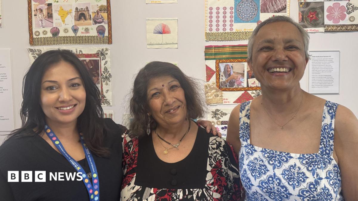 Three people stand close together indoors in front of a wall decorated with colourful artwork.