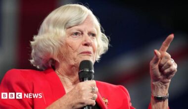 Ann Widdecombe wears a red jacket and holds a microphone while speaking at the Reform Party 2024 Conference.