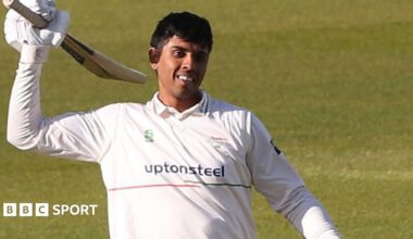Rishi Patel wearing cricket whites celebrates scoring a century by raising his bat