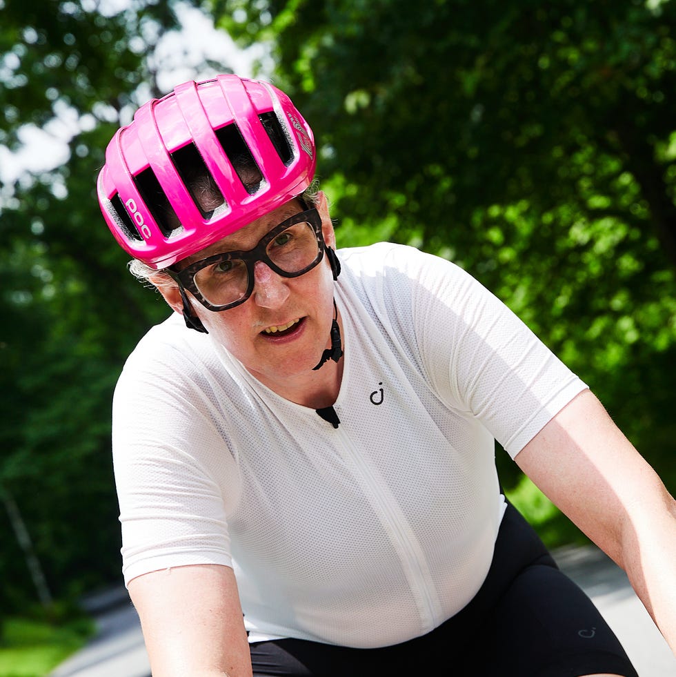 A cyclist wearing a pink helmet and glasses, riding on a tree-lined road during daytime.